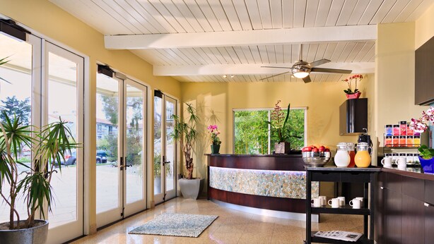 A reception area with a counter, potted plants, a coffee station, and a view through large glass doors. - The Inn at East Beach (Santa Barbara)