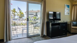 A hotel room with a bed, a TV, a desk, and a view of the street and palm trees outside. - The Inn at East Beach (Santa Barbara)