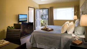 A hotel room with a bed, bedside table, lamp, and a television on a dresser. - The Inn at East Beach (Santa Barbara)