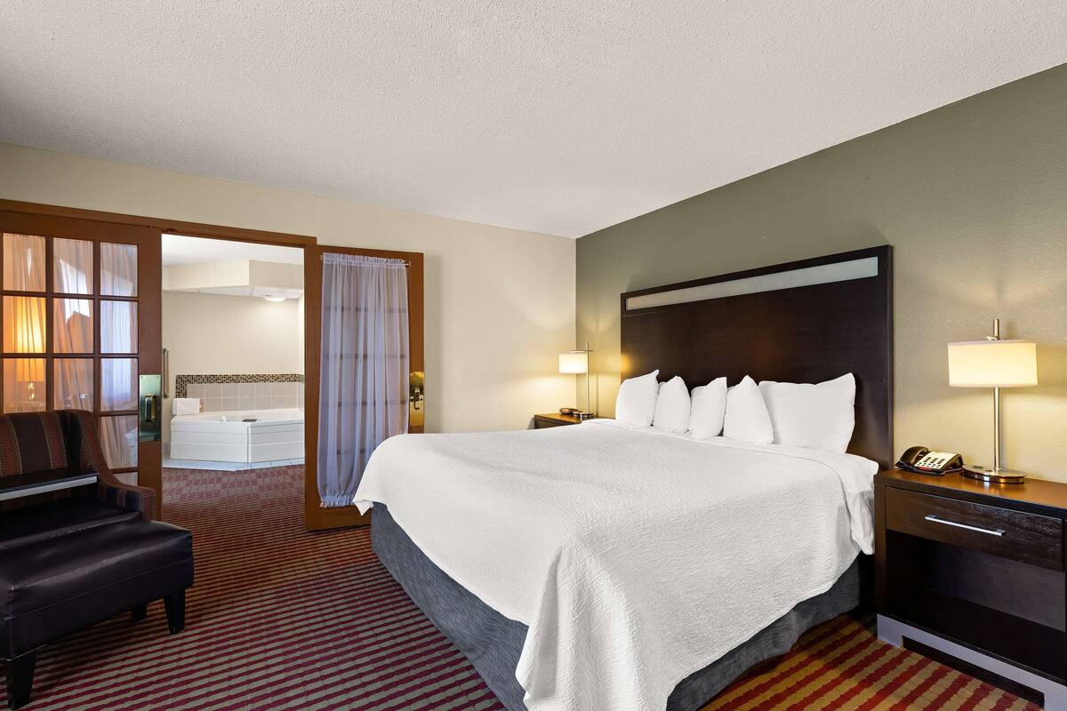 A hotel room with a large bed, a desk with a lamp, a chair, and a bathroom visible through an open door.