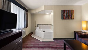 A hotel room with a flat-screen TV, a large window with curtains, a white bathtub, a colorful artwork on the wall, and a bedside table with a lamp. - Best Western Germantown Inn (Village of Germantown)