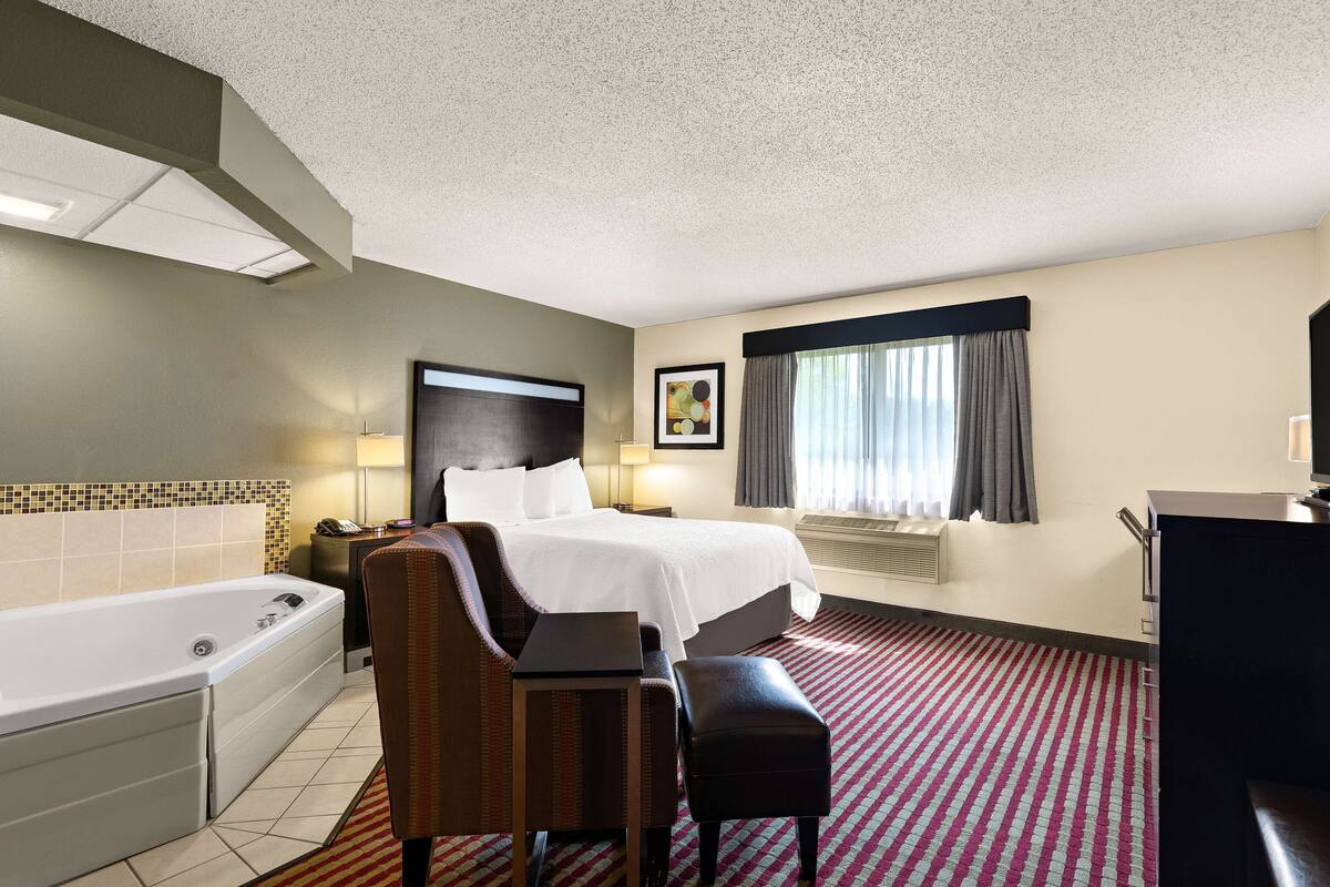 A hotel room with a bed, a desk, a chair, a desk lamp, a TV, a window with curtains, a bathroom with a bathtub, and a tiled wall.