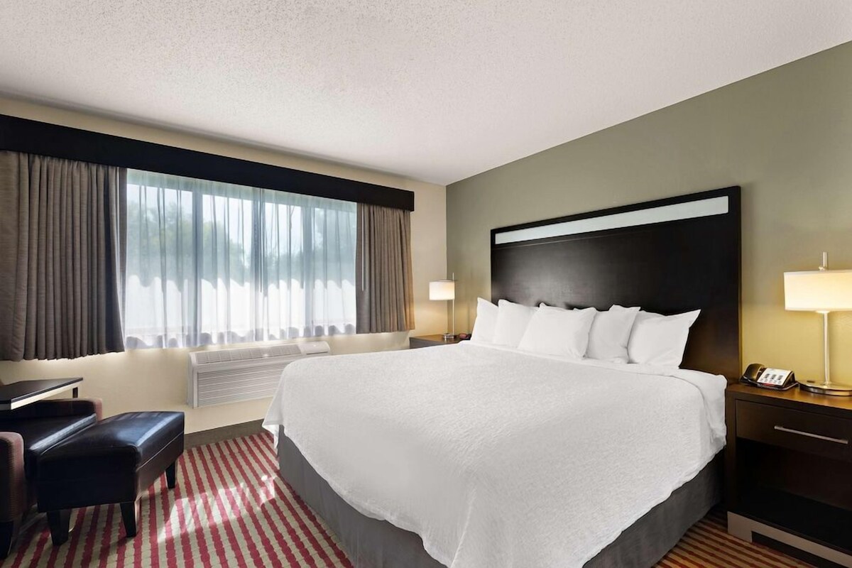 A hotel room with a large bed, a desk, a chair, a lamp, and a window with curtains.