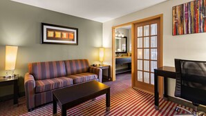 A hotel room with a striped sofa, a coffee table, a TV, a desk with a lamp, and a bathroom visible through a glass door. - Best Western Germantown Inn (Village of Germantown)