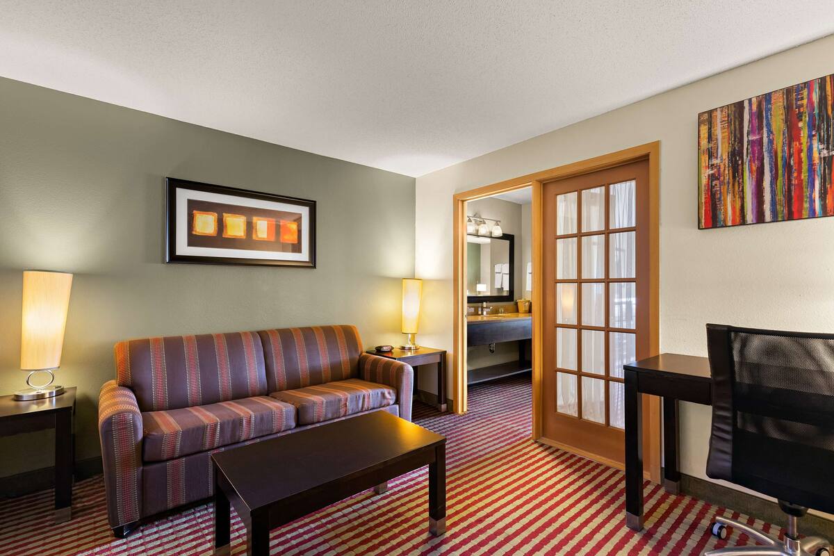 A hotel room with a striped sofa, a coffee table, a TV, a desk with a lamp, and a bathroom visible through a glass door.
