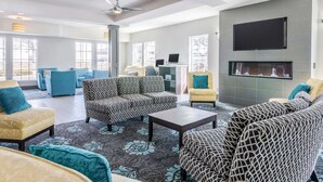 A spacious living area with a sectional sofa, armchairs, and a coffee table. There is a flat-screen TV mounted on the wall and a fireplace below it. The room has large windows allowing natural light and views of the outside. - La Quinta Inn & Suites by Wyndham Kansas City Airport (Kansas City)