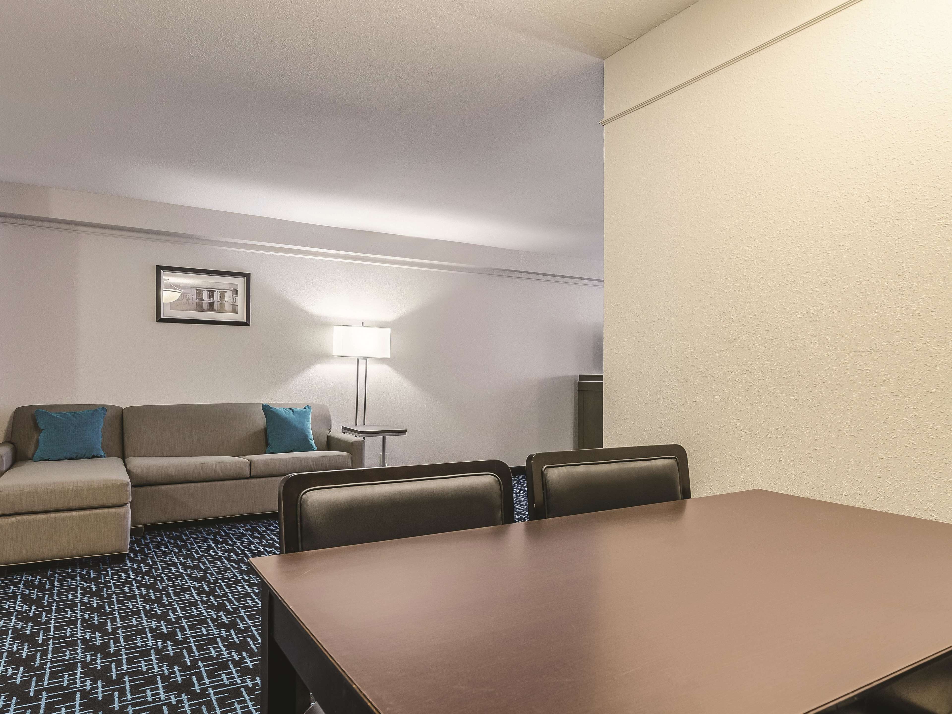 Hotelkamer met een grote vergadertafel, een bank met blauwe kussens, een vloerlamp en een ingelijst schilderij aan de muur.