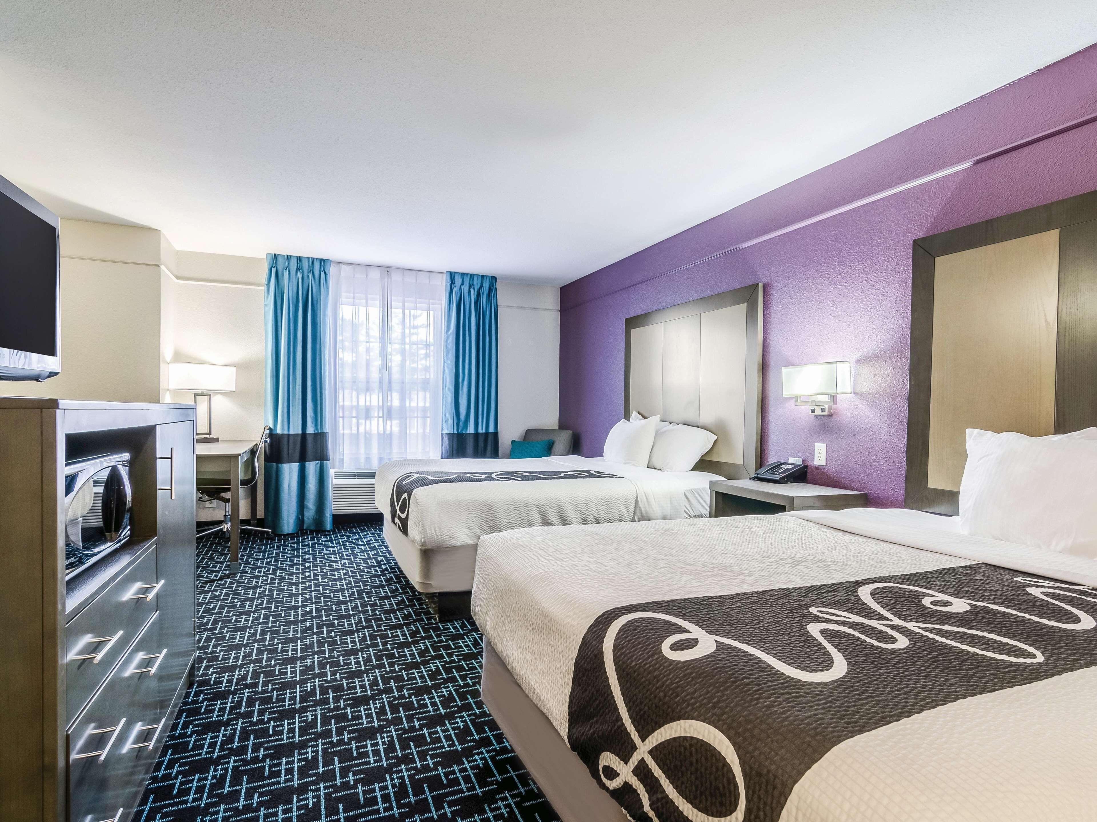 A hotel room with two beds, a TV, a desk, and a window with curtains.