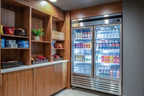 Een snackbar met houten planken vol met verschillende snacks en drankjes, en een grote koelkast gevuld met flessen water en frisdranken.