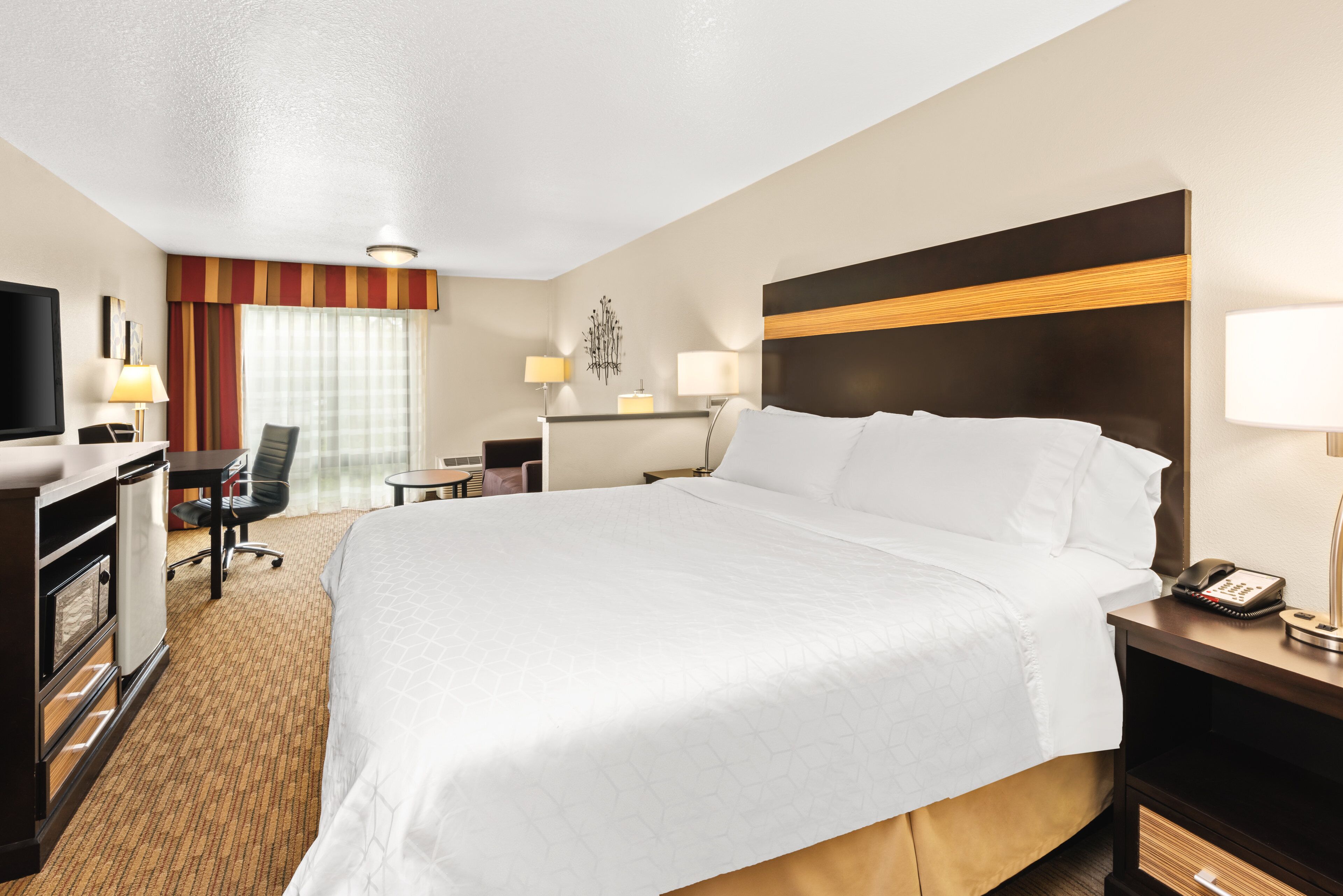 A hotel room with a large bed, a desk with a chair, a television, and a minibar.