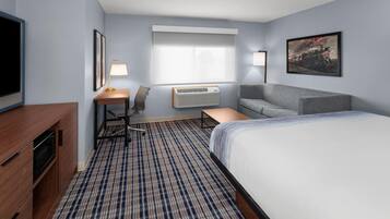 A hotel room with a bed, a desk, a chair, a TV, an air conditioner, and a painting on the wall.