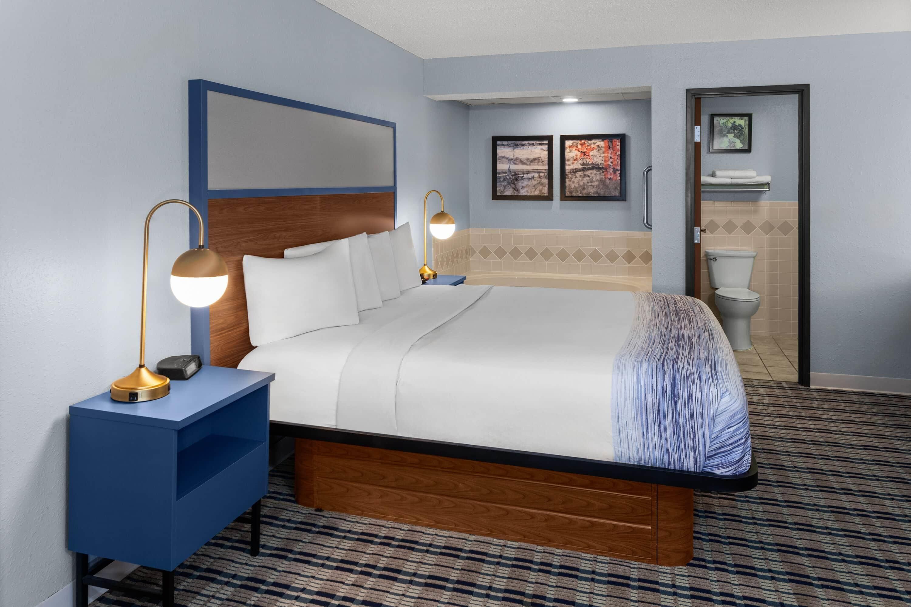 A hotel room with a bed, bedside lamps, a headboard, a wall-mounted mirror, a framed picture, a bathroom with a toilet and sink, and a patterned carpet.