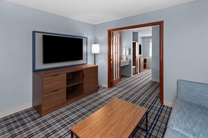 Habitación de hotel con una televisión de pantalla plana montada en la pared, un mueble de madera, una mesita pequeña, un sofá azul y una alfombra estampada.