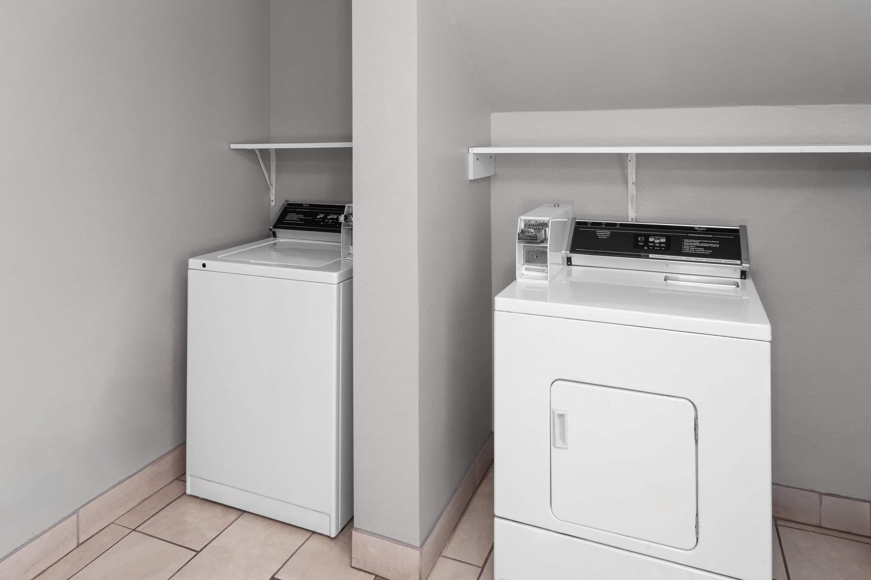Two white front-loading washing machines in a corner with a tiled floor and a white wall.