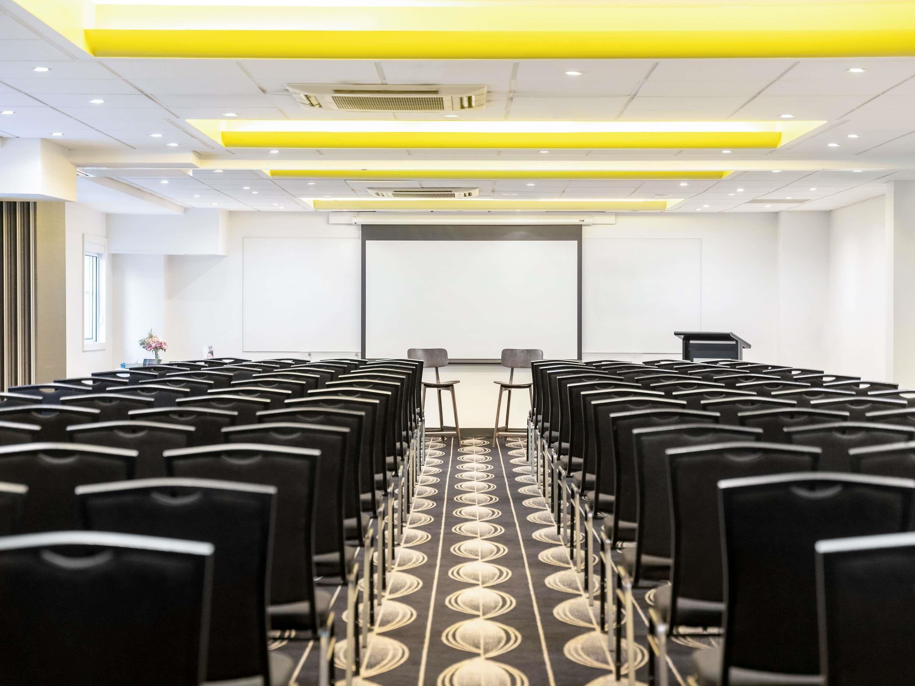 Ein Konferenzraum mit Reihen schwarzer Stühle, einer Projektionsleinwand und einem Präsentationstisch.