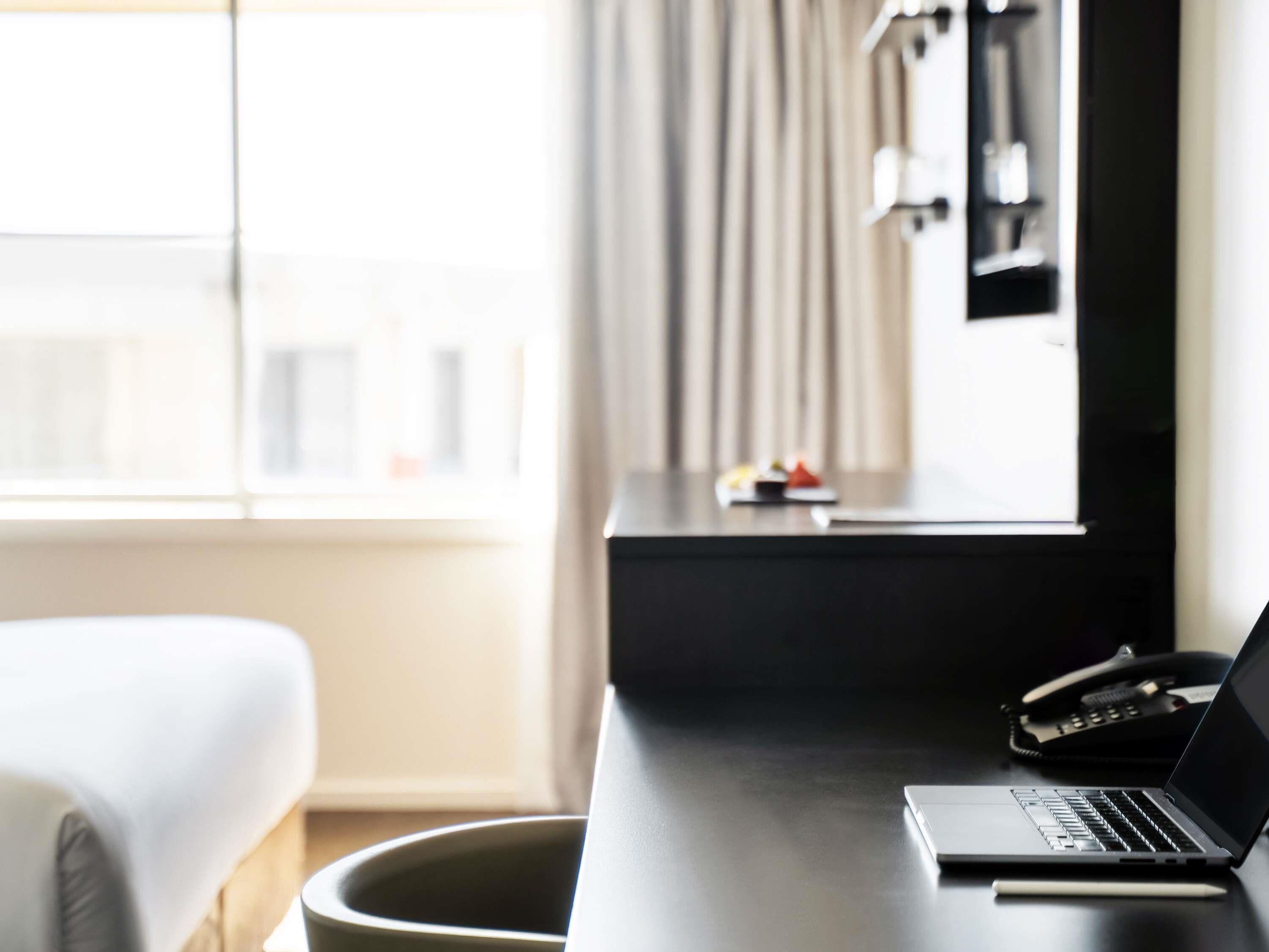 A hotel room with a bed, a desk with a laptop, a phone, and a window with curtains.
