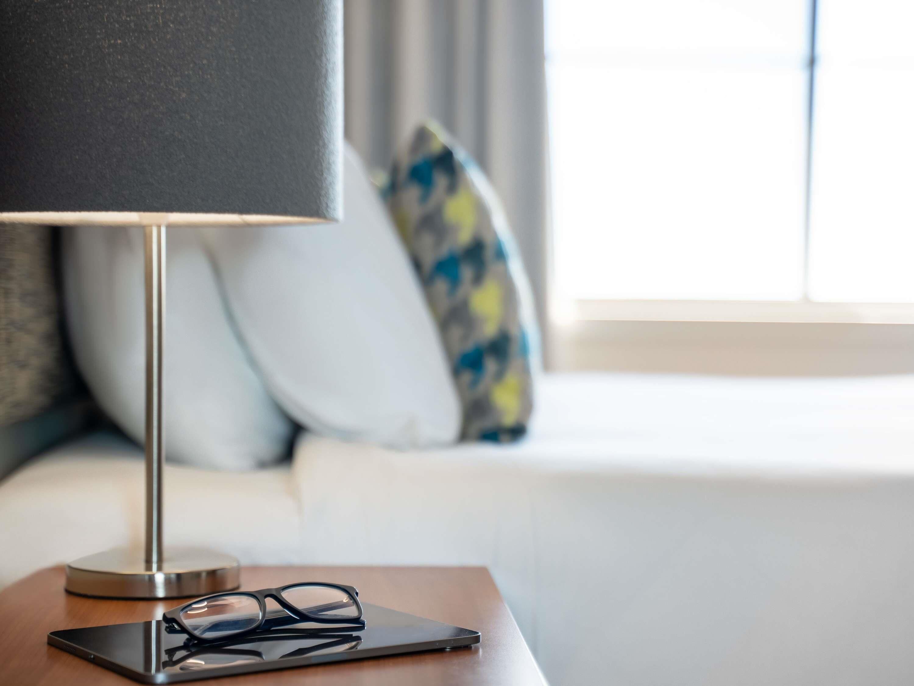 Een net opgemaakt bed met een witte dekbedovertrek en twee kussens, een nachtkastje met een lamp en glazen, en een raam met gordijnen.