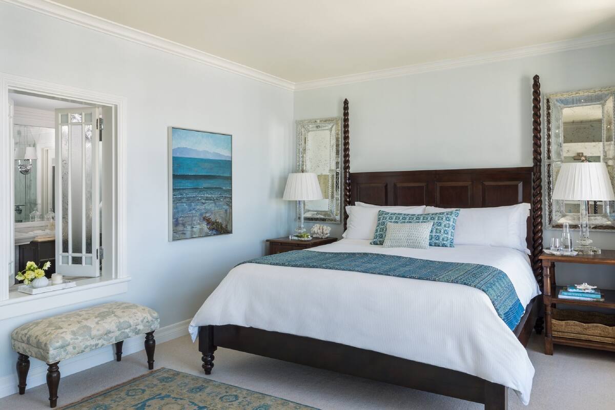 A bedroom with a large bed, two bedside tables with lamps, a bench, a painting, and a mirror.