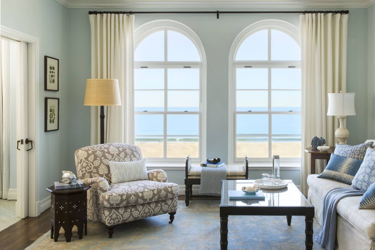 A room with a view of the beach, featuring a patterned armchair, a sofa with patterned cushions, a coffee table, and two lamps.