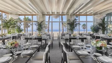 A dining area with tables set for service, large windows with a view of the ocean, and palm trees.