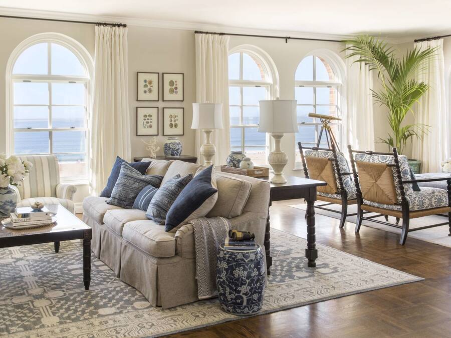 Uma sala de estar ampla com um sofá grande, poltronas e uma mesa de centro. Há várias janelas com vista para o oceano, e o ambiente é decorado com tapetes estampados e objetos decorativos.