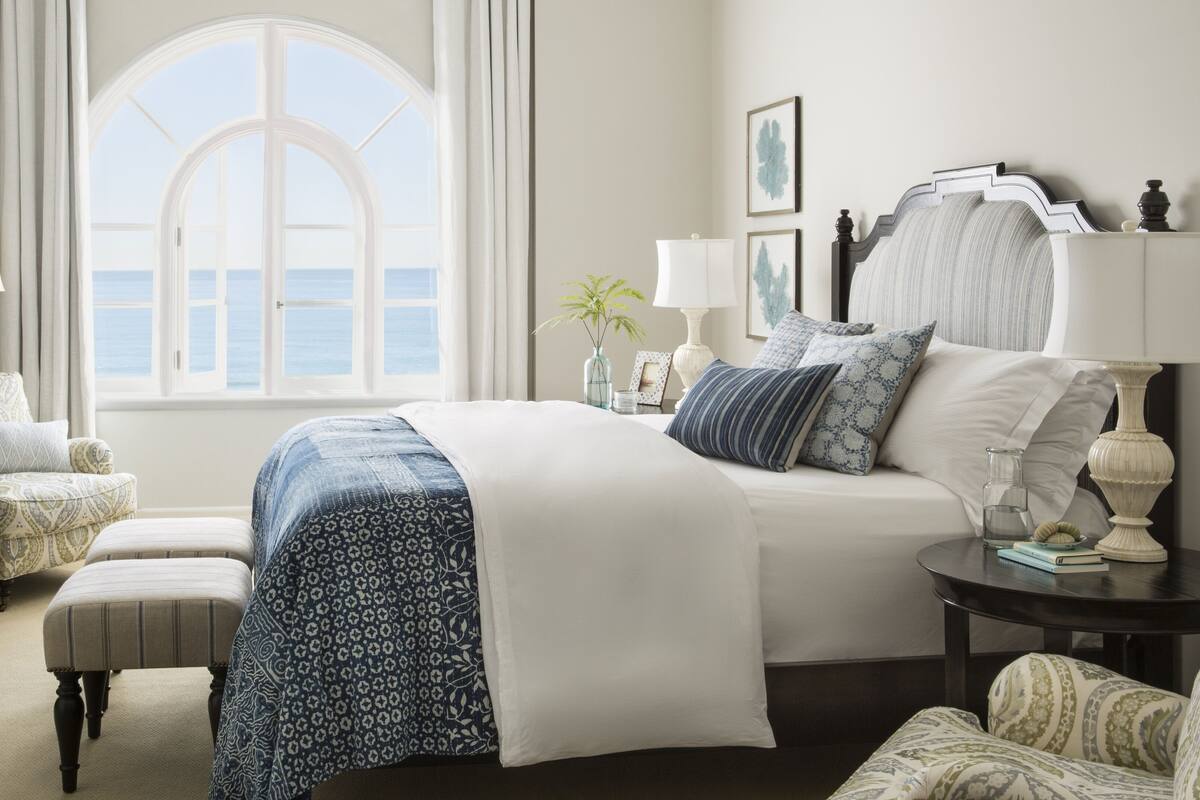 A bedroom with a large bed, a chair, a nightstand, and a view of the sea through a window.