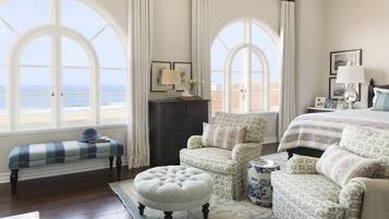 A bedroom with a large window overlooking the beach, a bed with a patterned bedspread, two armchairs, a bench, and a dresser with a lamp.