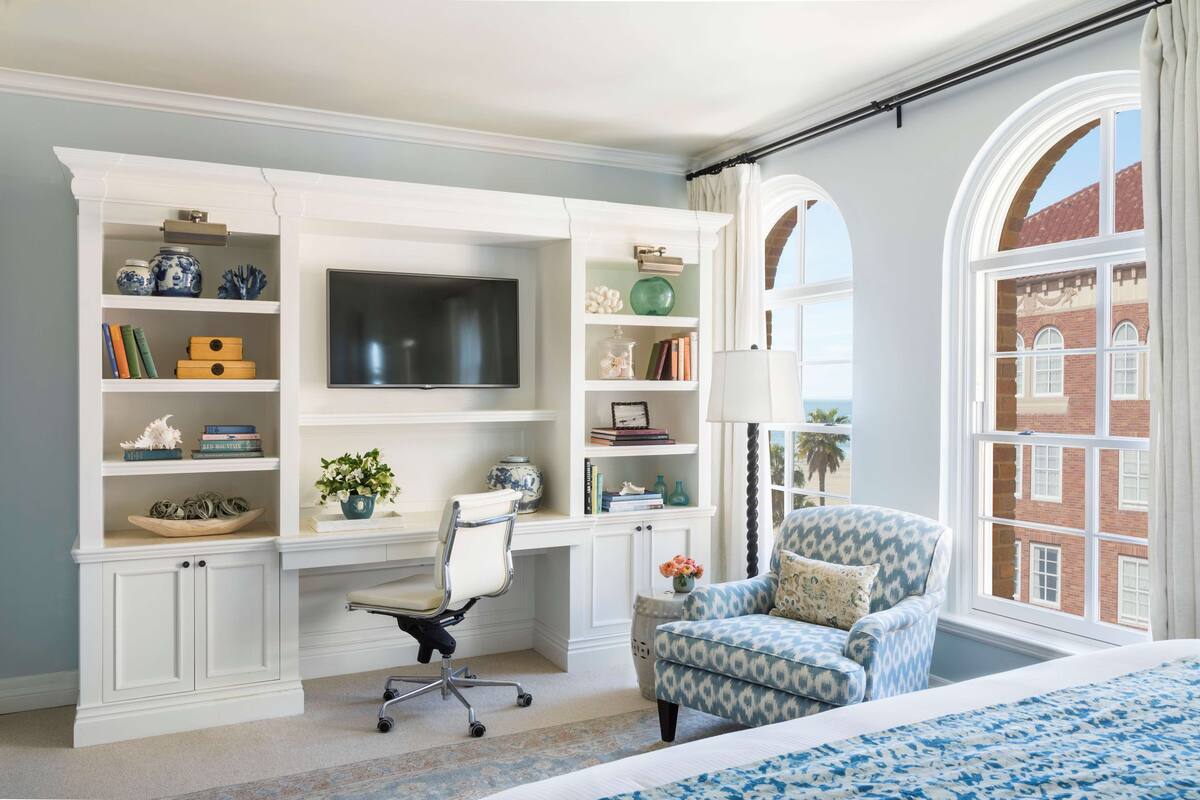 A bedroom with a large bed, a desk with a TV, a chair, and a window with a view of a building.