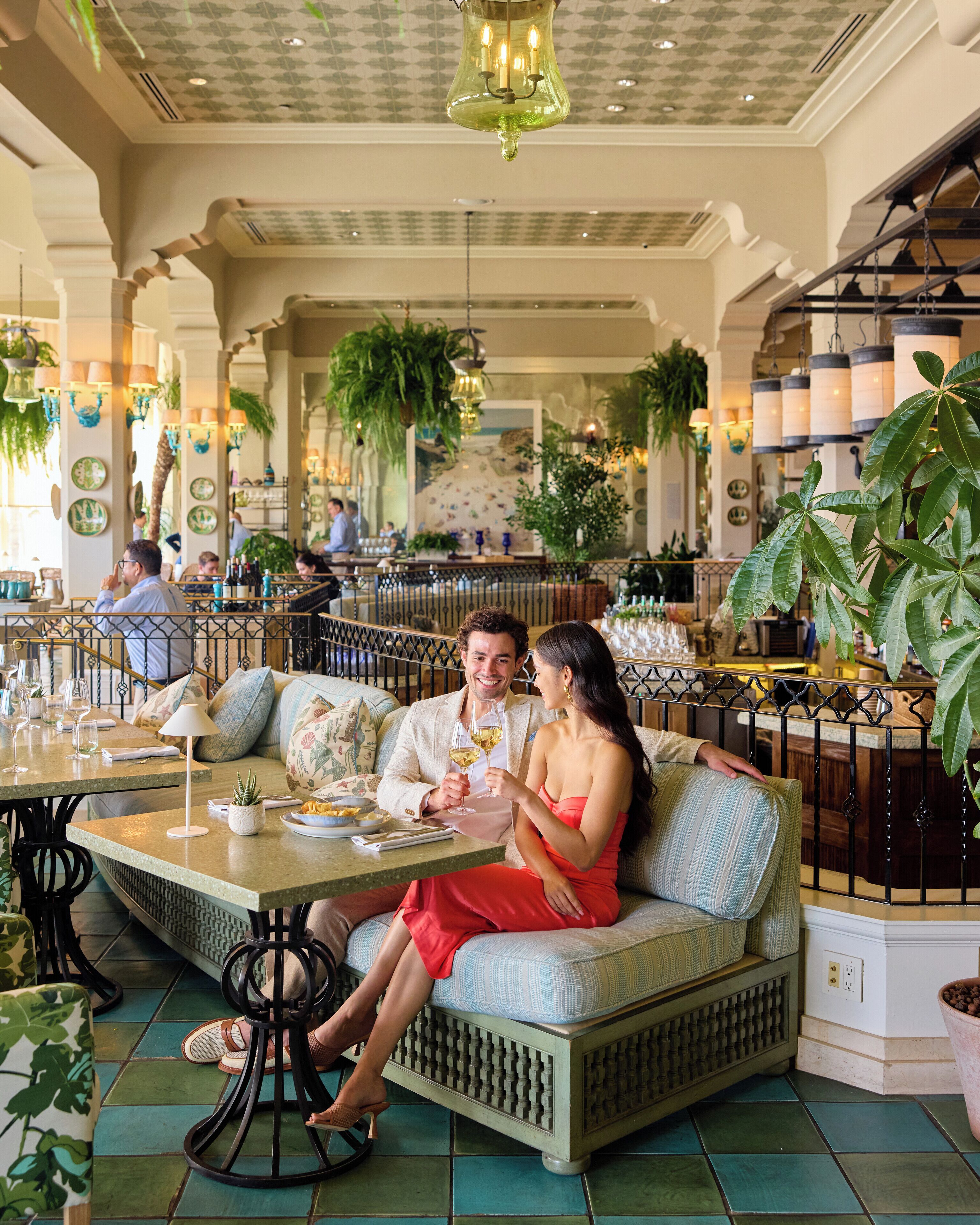 A couple seated at a table in a restaurant with a tiled floor, surrounded by greenery and other diners.