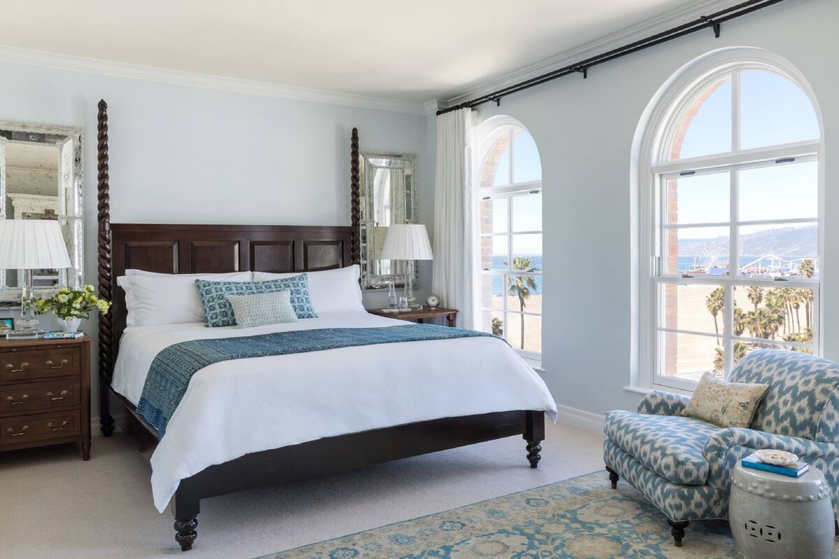 A spacious bedroom with a large bed, two bedside tables, a nightstand, a chair, and a view of the beach.