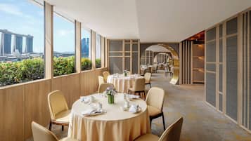 Habitación de hotel con vista a un horizonte urbano, un área de comedor con mesas redondas y un mostrador de recepción.