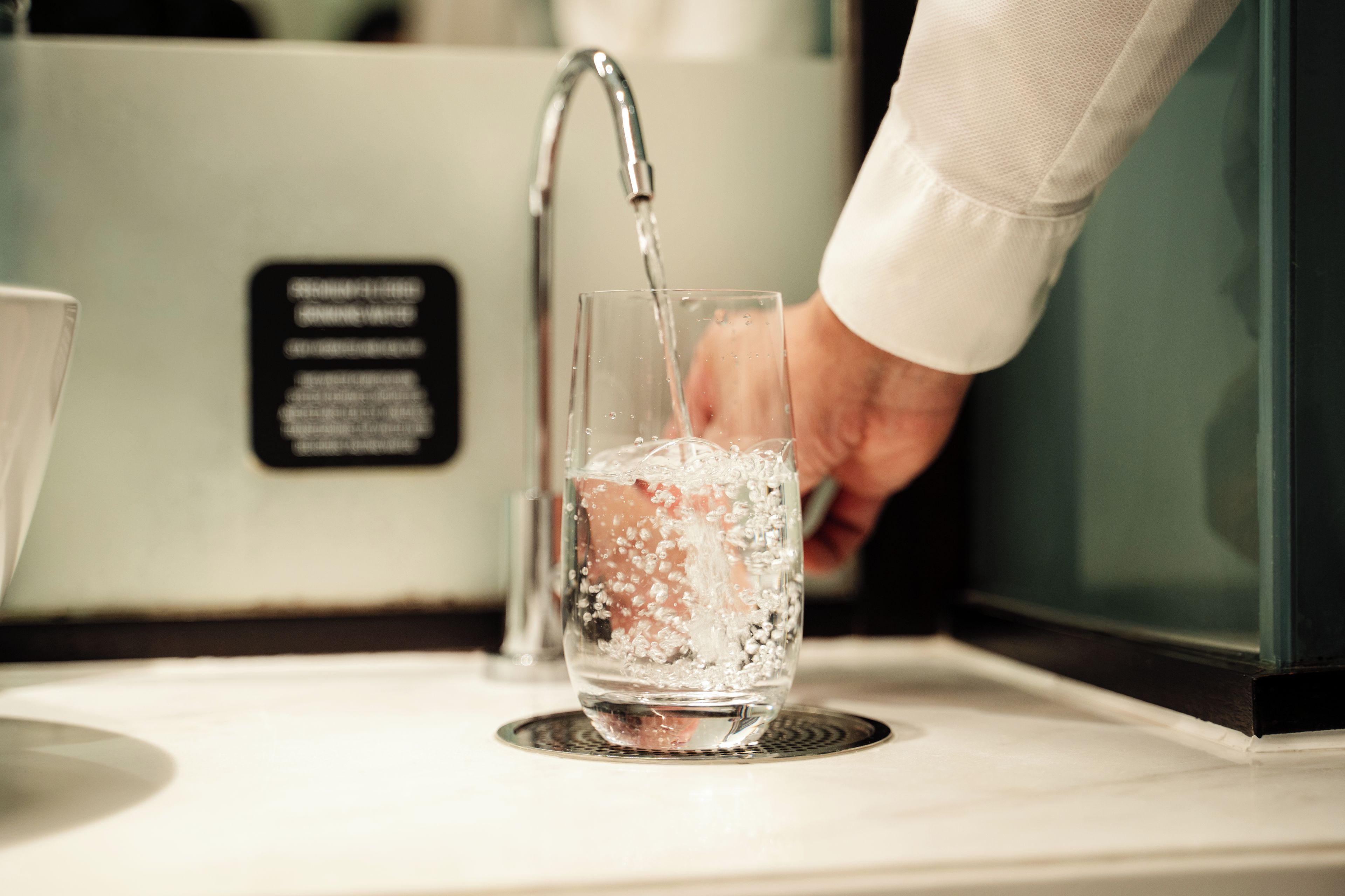 Una mano vertiendo agua en un vaso con burbujas, un grifo y un cartel con texto.
