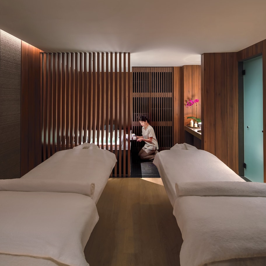 A spa room with two massage tables, a wooden partition, and a small shelf with a vase of flowers.