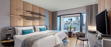 A modern hotel room with a large bed, a desk, a flat-screen TV, and a view of a cityscape through the window.