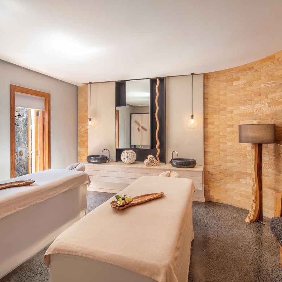 A spa room with two massage tables, a stone wall, a wooden wall, a large mirror, and a wooden floor.