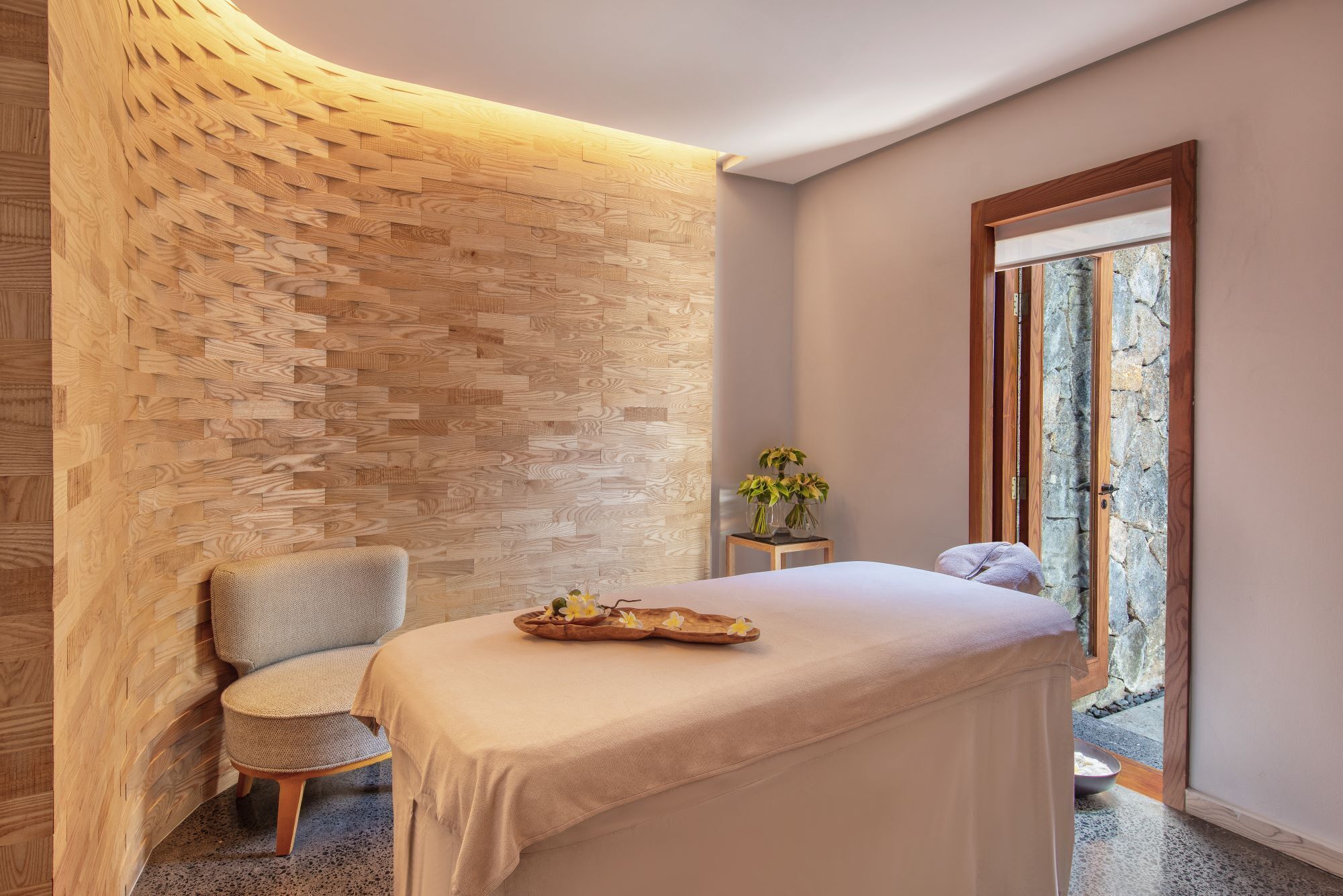 A spa room with a wooden wall, a massage table, a chair, and a small table with flowers.