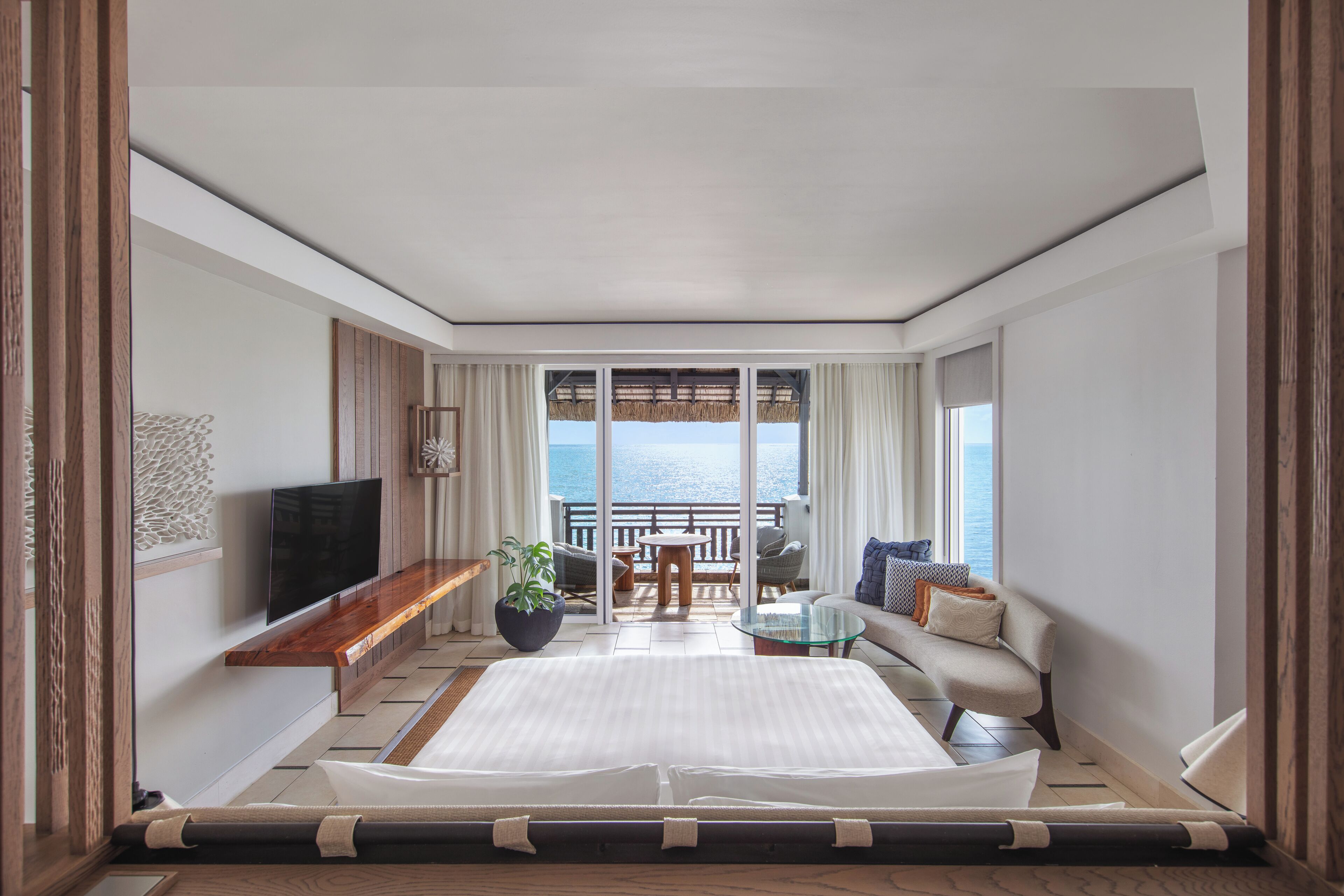 A hotel room with a bed, a TV, a balcony with a view of the sea, a seating area with a table, and a potted plant.