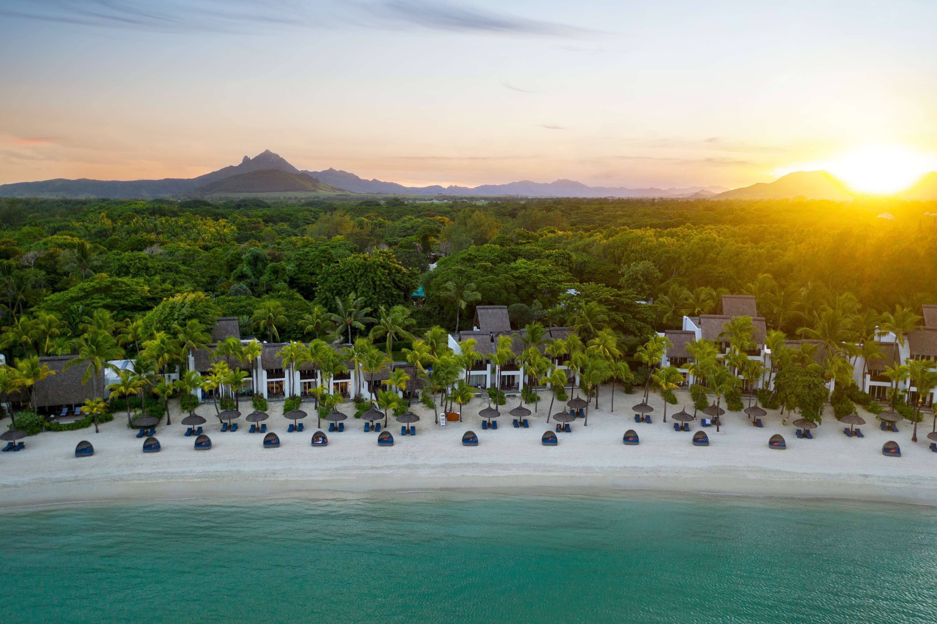 Un resort en la playa con cabañas de techo de paja, palmeras y una montaña al fondo.