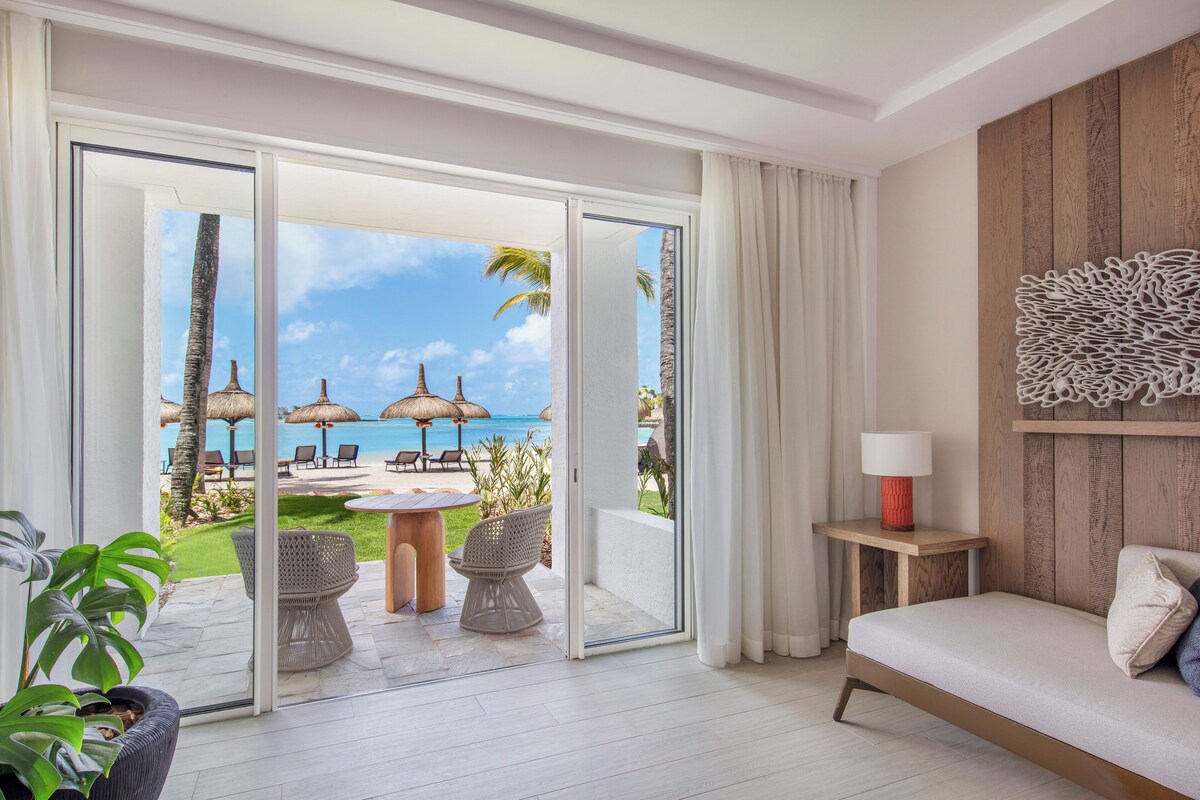 Ein Zimmer mit einem Bett, einem Tisch, Stühlen und Blick auf einen Strand mit Sonnenschirmen sowie einer Holzwand.