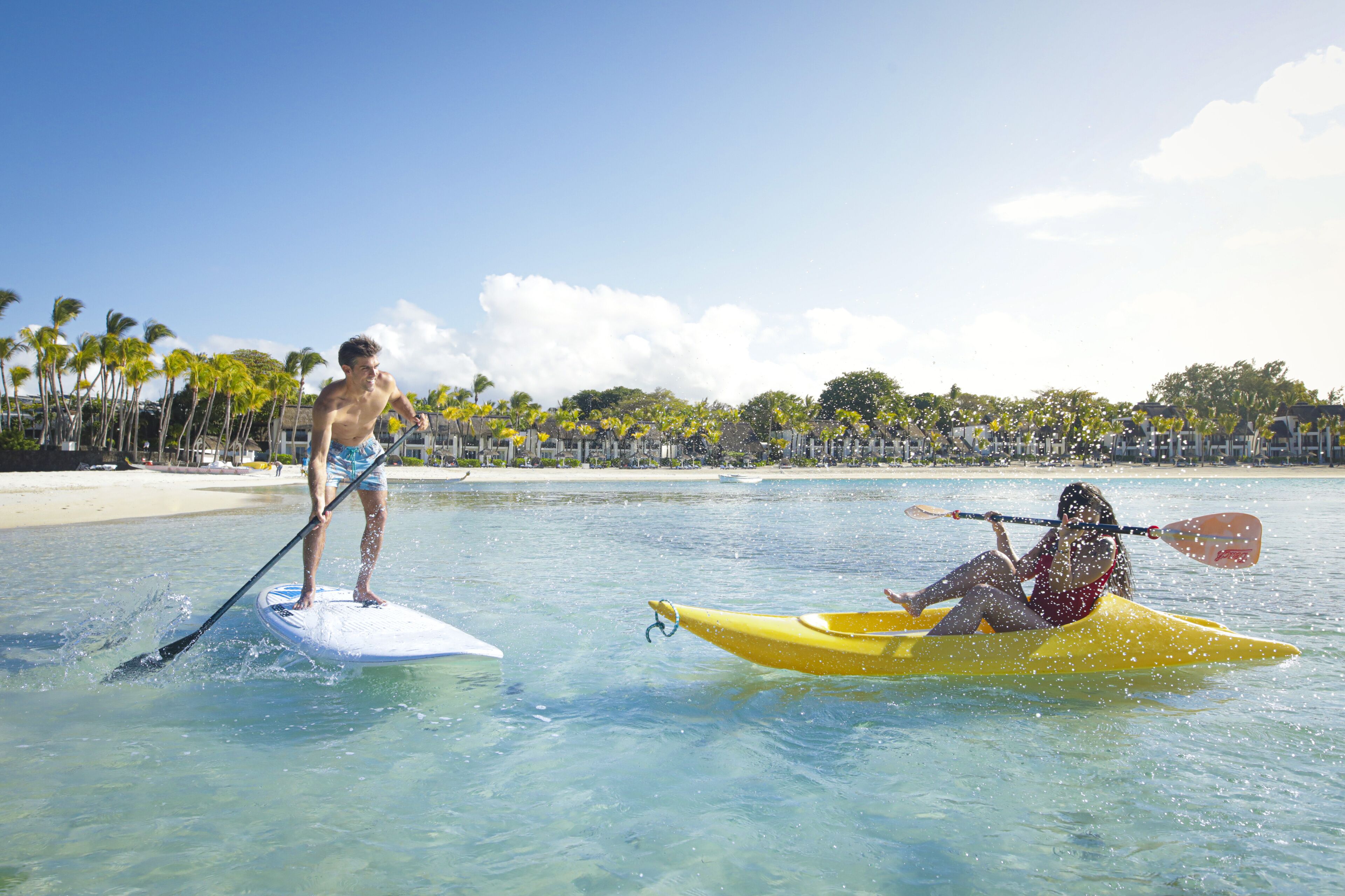 A man standing on a paddleboard and a woman in a kayak on a beach with palm trees and a clear sky.