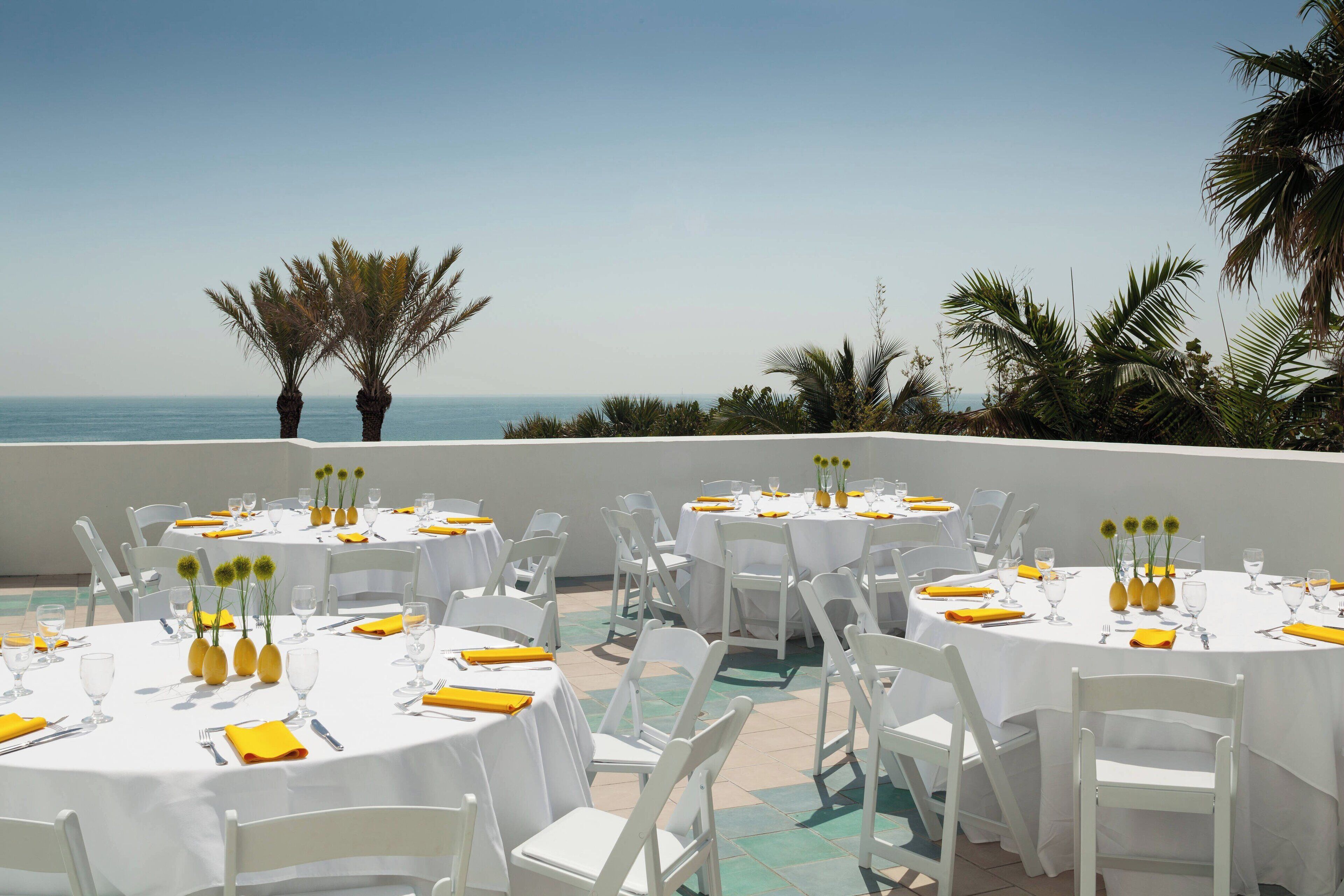 Un área de comedor al aire libre con mesas redondas, sillas blancas y servilletas amarillas.