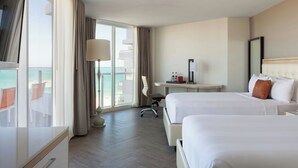 Ett hotellrum med två sängar, ett skrivbord, en stol och ett stort fönster med utsikt över stranden.