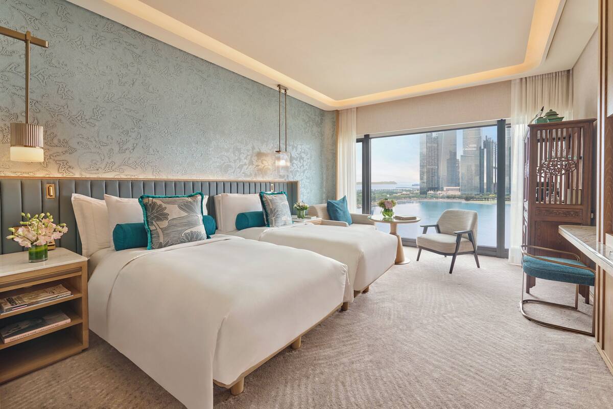 Habitación de hotel con dos camas, un escritorio, una silla y un amplio ventanal con vista a la ciudad.
