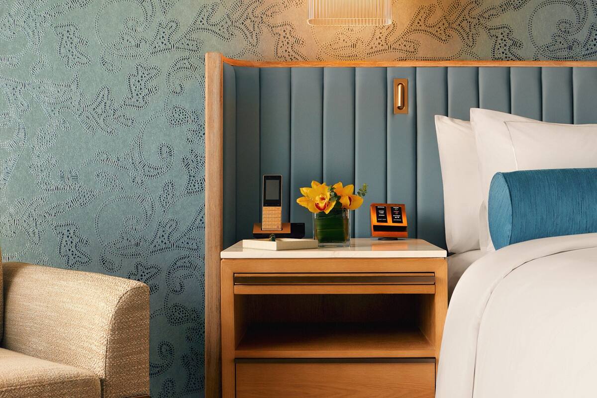 Een netjes opgemaakte slaapkamer met een houten nachtkastje, een bijzettafeltje, een bed met wit- en blauwe beddengoed, en een klein tafeltje met een vaas geel bloemen.