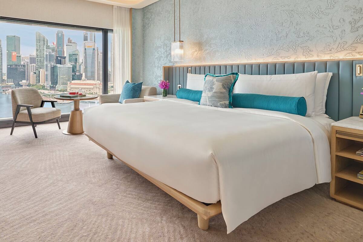Een moderne hotelkamer met een groot bed, uitzicht op de stadshemel, een raam met stadszicht, een zitje met stadszicht, een nachtkastje met een bloemenvaas en een bedlampje.