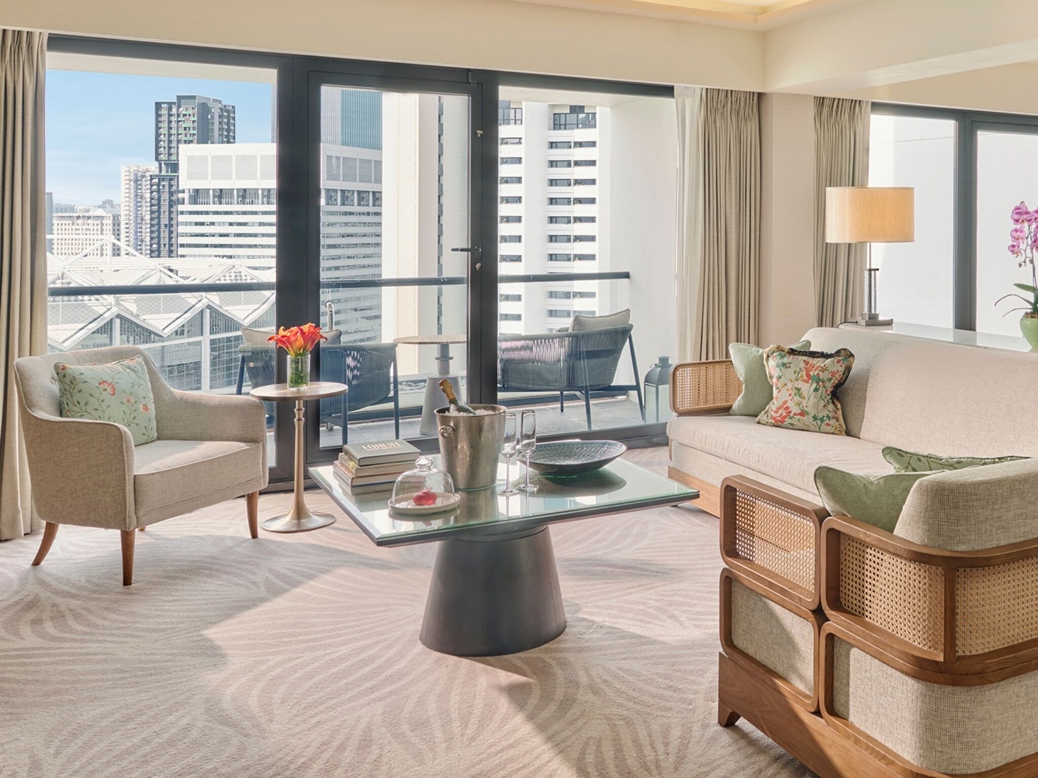 A modern hotel room with a large balcony, a sofa, a chair, a coffee table, and a view of the cityscape.