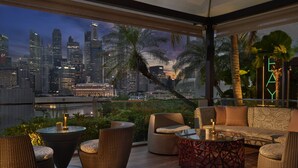 Un salón en la azotea con mobiliario de mimbre, una mesa de vidrio y vistas a un horizonte urbano al anochecer.