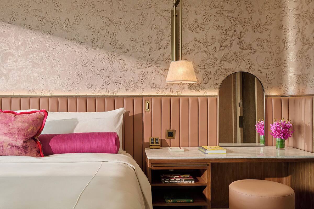 Habitación de hotel con cama, mesita de noche, espejo y escritorio con lámpara.