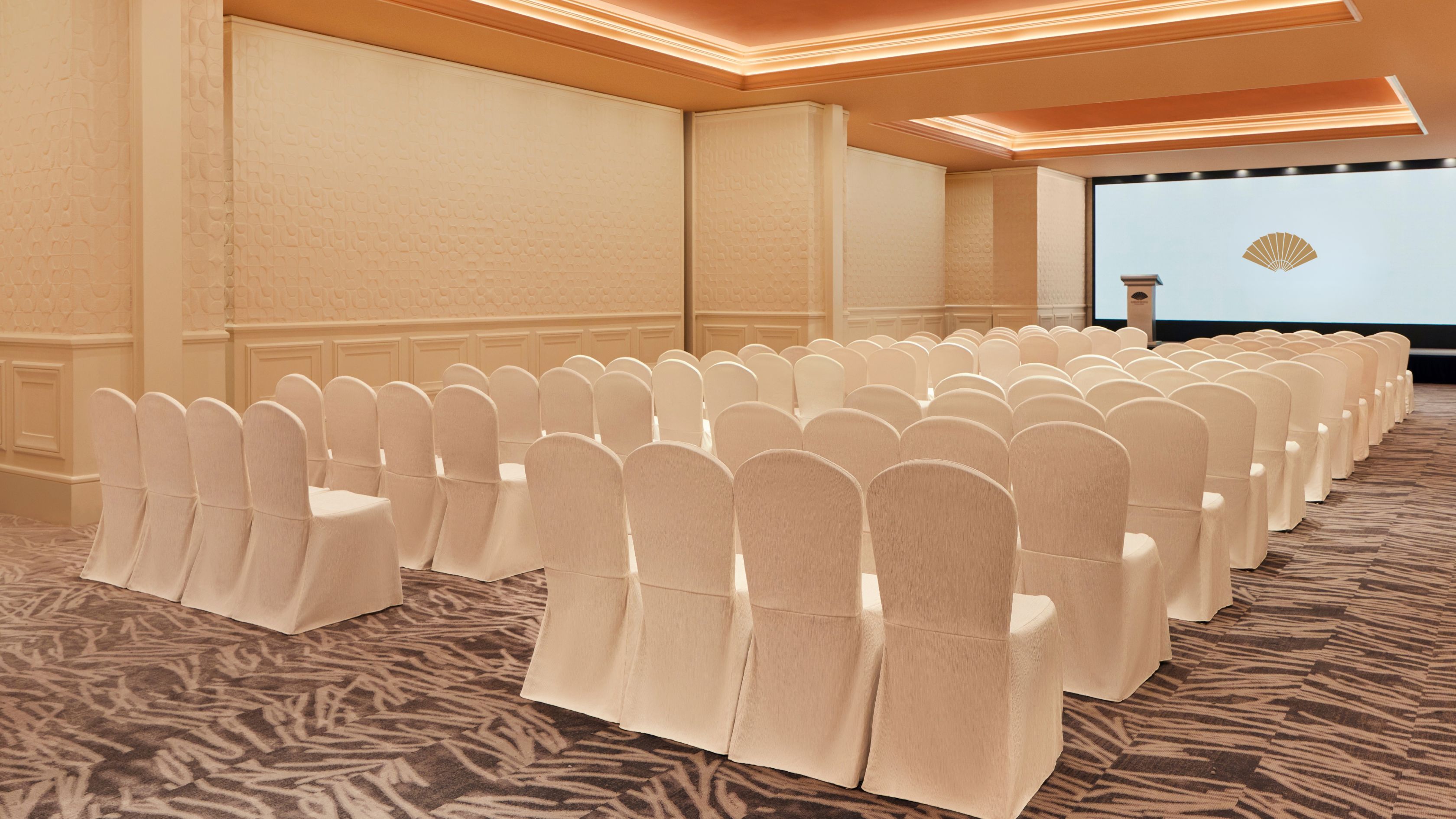 Un salón de conferencias con filas de sillas cubiertas con fundas blancas, suelo alfombrado y una pantalla grande al frente.