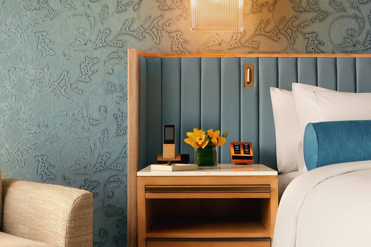 Un dormitorio ordenado con una mesita de noche de madera, una mesilla auxiliar, una cama con ropa de cama blanca y azul, y una pequeña mesa con un jarrón de flores amarillas.
